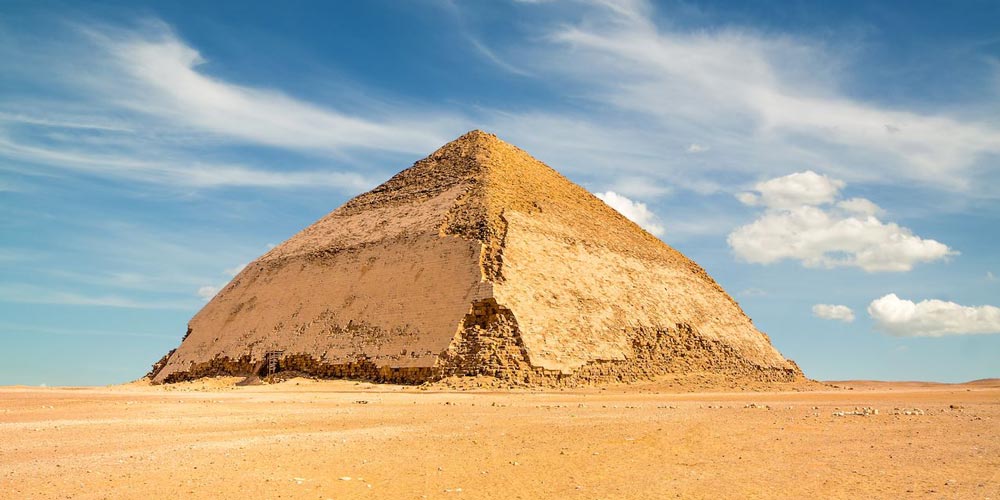 Ein atemberaubender Blick auf die Knickpyramide in Dahshur, Ägypten, mit ihrer einzigartigen schrägen Struktur unter einem Himmel voller fluffiger Wolken, die die architektonische Genialität des alten Ägypten hervorhebt.
